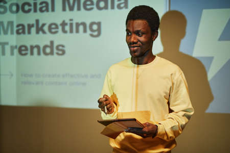 Waist up portrait of young black man giving presentation on marketing while standing by projector screen and addressing audience, copy spaceの写真素材
