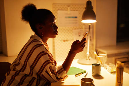 Side view portrait of young black businesswoman working at night in cozy yellow lighting and checking smartphone messagesの写真素材