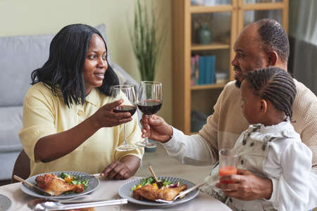 African couple toasting with glasses of red wine while having dinner with child together at dining tableの写真素材