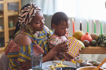 African woman in national costume opening present with her little daughter while they sitting at dining tableの写真素材