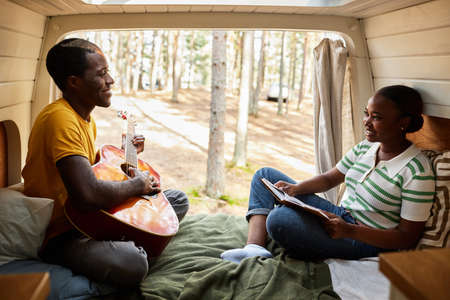 African young couple resting in van playing guitar and reading a book during camping in the forest