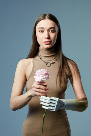 Portrait of young girl with prosthetic arm holding flower and looking at camera against blue backgroundの写真素材
