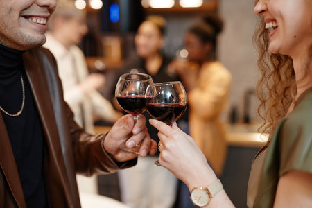 Close-up of hands of young smiling couple with red wine making toast and clinking with wineglasses afainst group of their friendsの写真素材