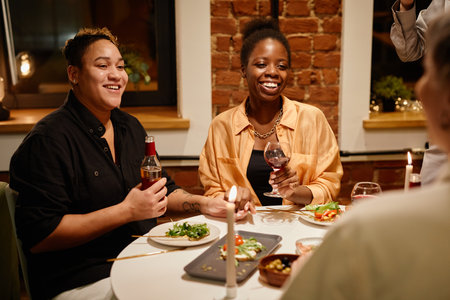 Happy young lesbian couple with alcoholic drinks sitting by served table and talking to friends who...