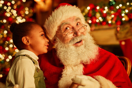 Portrait of cute black girl whispering secrets to Santa Claus on Christmas eveの写真素材