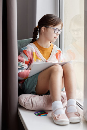 Vertical full length portrait of teenage girl with Down syndrome drawing pictures while sitting by...