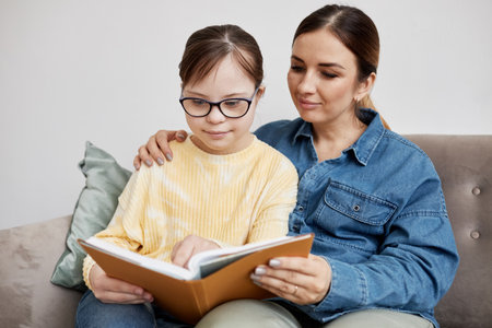 Portrait of teen girl with Down syndrome reading book with loving mother at homeの写真素材