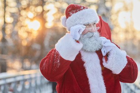 Waist up portrait of traditional Santa Claus carrying sack with presents in winter setting outdoors and looking up over eyeglasses, copy spaceの写真素材