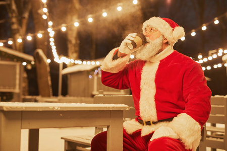 Portrait of traditional Santa Claus drinking hot cocoa in park with fairy lights in background, copy spaceの写真素材