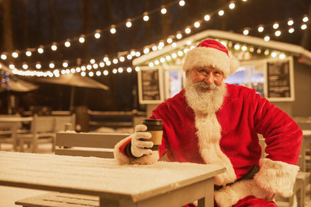 Portrait of traditional Santa Claus looking at camera with hot coffee cup in park with fairy lights in background, copy spaceの写真素材