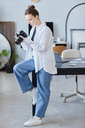 Vertical full length portrait of female photographer checking images in camera at studio minimal