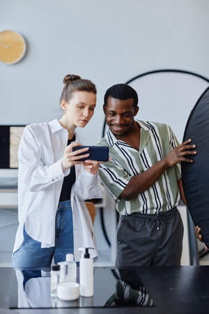 Vertical portrait of two young photographers working on product photography in studio and using mobile phone for social mediaの写真素材