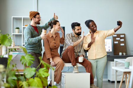 Side view of four young cheerful employees waving hands and looking at smartphone camera with smiles...
