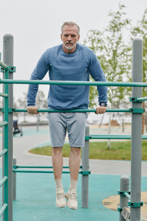Full length portrait of sportive mature man exercising on parallel bars outdoors and looking at...