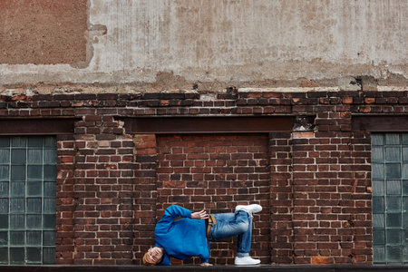 Wide angle shot of young man doing handstand breakdance poses outdoors against shabby brick wall,...
