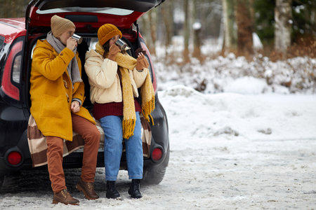 Full length portrait of young couple sitting in car trunk in winter forest while enjoying hot cocoa, copy spaceの写真素材
