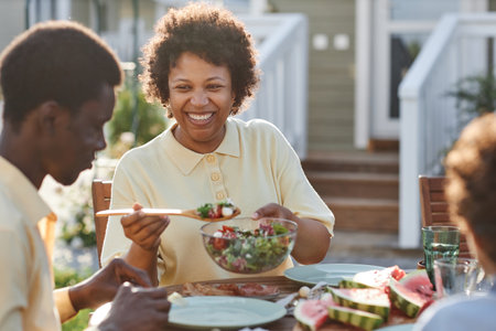 Portrait of smiling African American woman serving food to family while enjoying dinner together outdoors in sunlightの写真素材