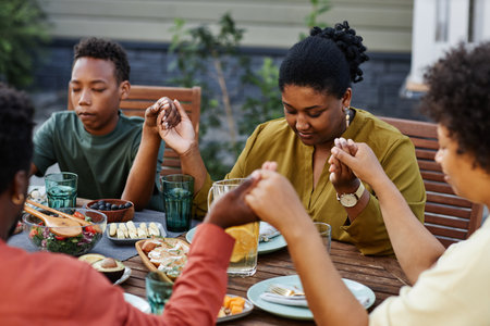 Portrait of black young woman saying grace at table outdoors during family gathering and holding handsの写真素材