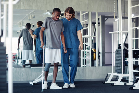 African patient trying to walk with his pain leg using walking stick with the help of doctor in gym
