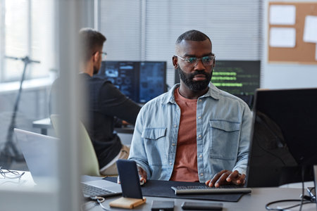 Portrait of black man using computer while programming mobile software in office behind glass wallの写真素材