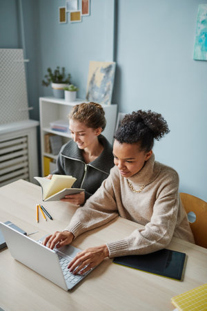 High angle view of young woman working on laptop with online project while her colleague reading...