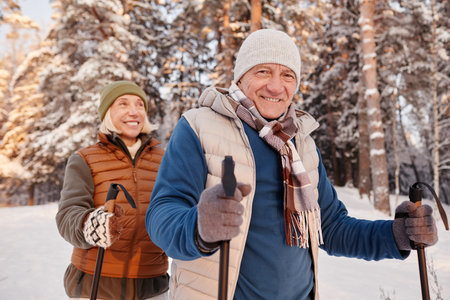 Waist up portrait of happy senior couple walking with poles in winter forest and smiling at cameraの写真素材
