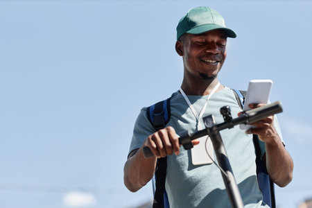 Low angle portrait of young black boy using smartphone while riding electric scooter against blue sky, copy spaceの写真素材