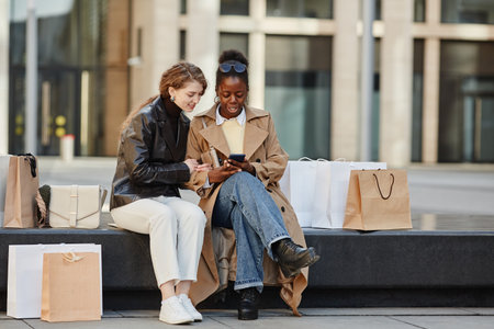 Full length portrait of two young women using smartphone while relaxing in downtown city after shopping dayの写真素材