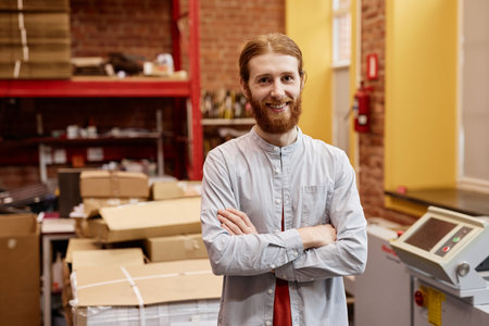 Waist up portrait of smiling young man working in industrial printing shop and standing with arms crossed, copy spaceの写真素材