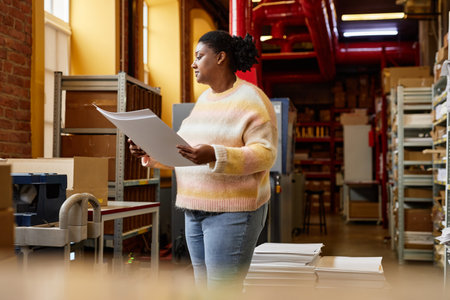 Side view portrait of black young woman holding prints by industrial printing machine at workshop,...