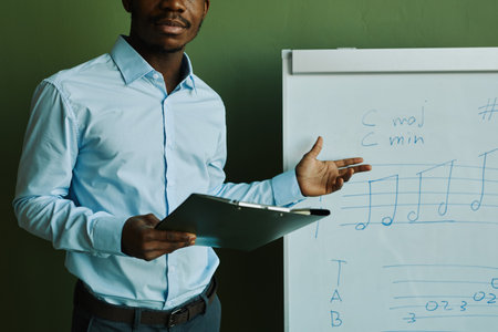 Young African American man in blue shirt standing by whiteboard with musical notes and explaining them to audience at lessonの写真素材