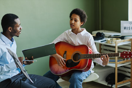 Youthful African American schoolgirl playing acoustic guitar at lesson in music school while sitting in front of teacher consulting herの写真素材