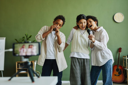 Two intercultural girls in casualwear singing in microphone while African American boy playing...
