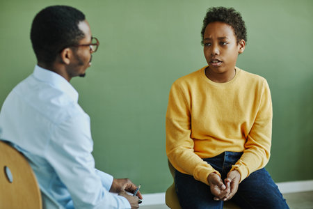 Upset schoolboy in casualwear describing his problem to young African American school psychologist during individual session after classesの写真素材