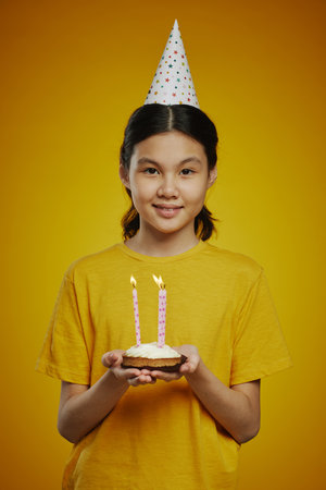 Smiling youthful girl in birthday cap and yellow t-shirt holding small homemade cake with several...