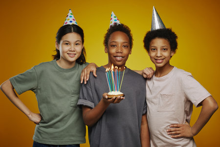Three youthful multicultural friends in birthday caps and casualwear standing in front of camera while boy holding cake with candlesの写真素材