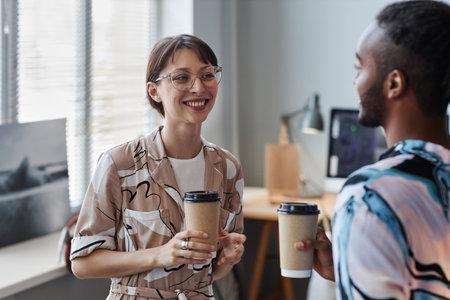Waist up portrait of smiling young woman enjoying takeout coffee while chatting with coworker at workplaceの写真素材
