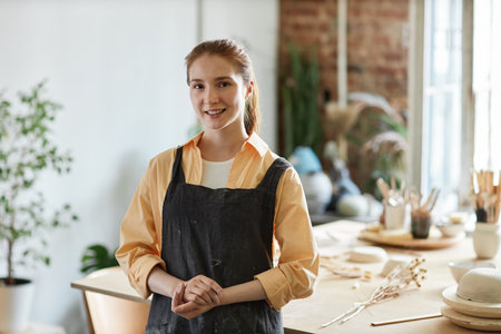 Waist up portrait of smiling young woman wearing apron while standing in pottery studio and looking...