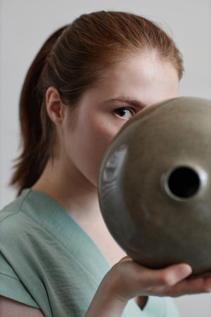 Close up of young woman holding handmade ceramic vase to face and looking at cameraの写真素材