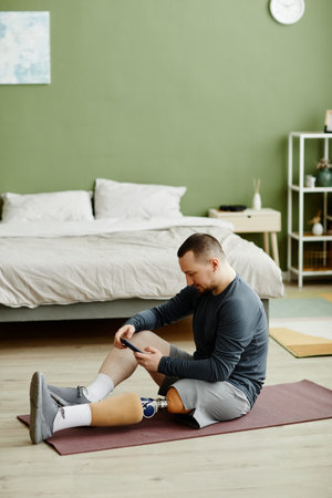 Vertical side view portrait of adult man with prosthetic leg sitting on yoga mat at home and using smartphoneの写真素材