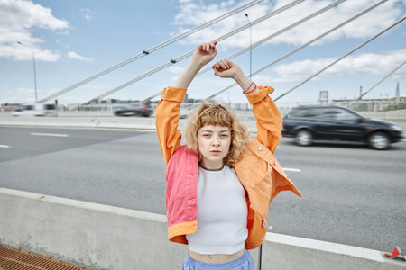 Waist up portrait of free young woman wearing colorful clothes on city bridge and looking at camera, copy spaceの写真素材