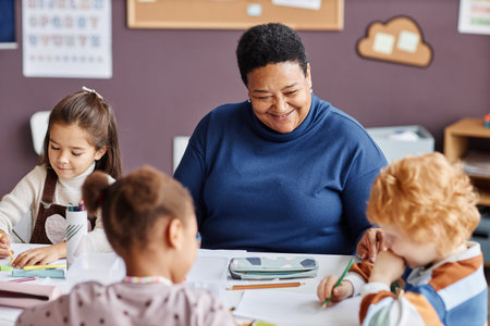 Focus on happy African American mature teacher and little learner with dark long hair sitting in front of two intercultural nursery schoolkidsの写真素材