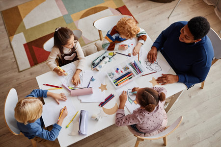 Above shot of several intercultural learners of nursery school and their teacher sitting in front of them during lesson of drawingの写真素材