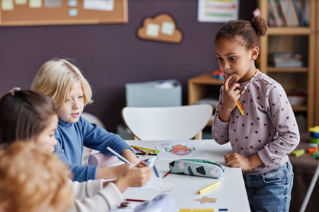 Adorable African American little girl with crayons looking at group of classmates sitting in front of her while thinking of new ideasの写真素材
