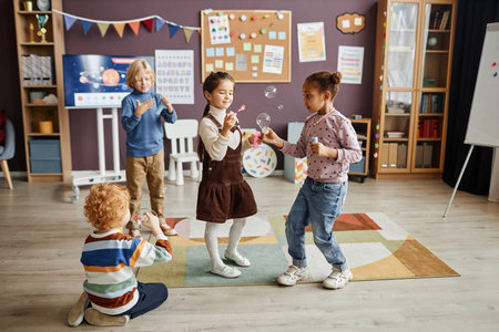 Group of little carefree kids in casualwear having fun with soap bubbles while standing on carpet in...