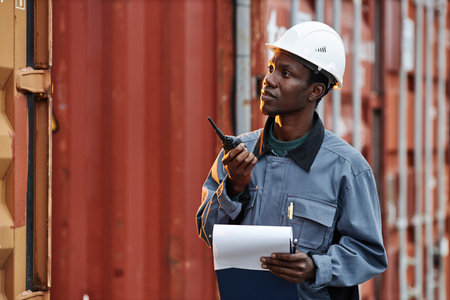 Waist up portrait of young black man using walkie talkie while working at shipping docks with...