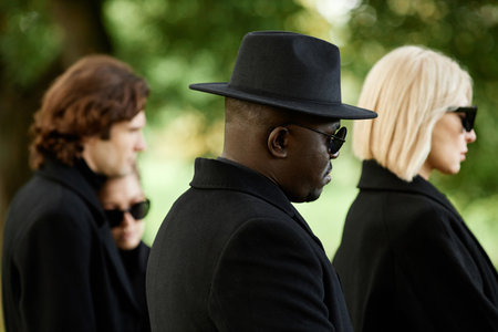 Side view of people wearing all black with sunglasses standing in silence at outdoor funeral...