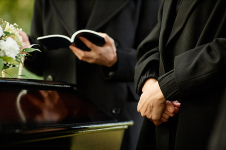 Close up of young woman standing by coffin at outdoor funeral ceremony, priest reading prayer in...