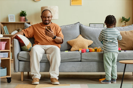 Full length portrait of father using smartphone with son playing on sofa at home, copy spaceの写真素材