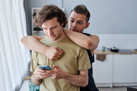 Cozy portrait of young same sex couple embracing in kitchen at home, man using smartphoneの写真素材